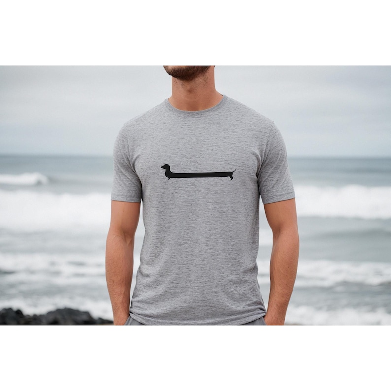 May include: A heather gray t-shirt featuring a black dachshund dog graphic. The dog is elongated, with a long body and short legs. The shirt is short-sleeved and has a crew neck. The background shows a beach scene with the ocean and sky.