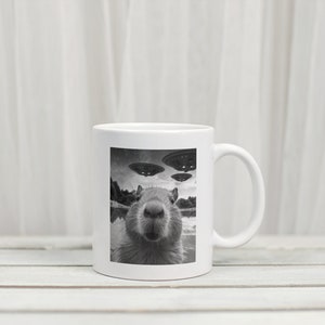 Pu&ograve; includere: Una tazza in ceramica bianca con un'immagine in bianco e nero di un capibara che guarda direttamente la fotocamera. Il capibara &egrave; in un specchio d'acqua con tre dischi volanti sullo sfondo.