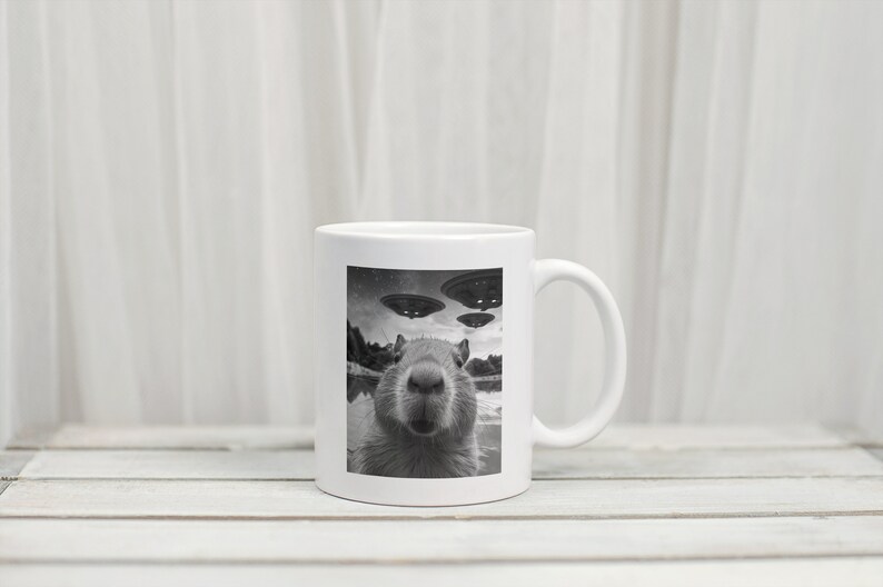 Pu&ograve; includere: Una tazza in ceramica bianca con un'immagine in bianco e nero di un capibara che guarda direttamente la fotocamera. Il capibara &egrave; in un specchio d'acqua con tre dischi volanti sullo sfondo.