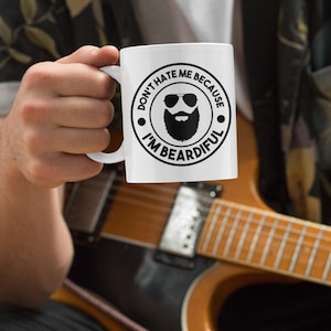 May include: White ceramic mug featuring the text "DON'T HATE ME BECAUSE I'M BEARDIFUL" in a black circular design. The design includes a beard, sunglasses, and the text. The mug is held in front of a guitar.