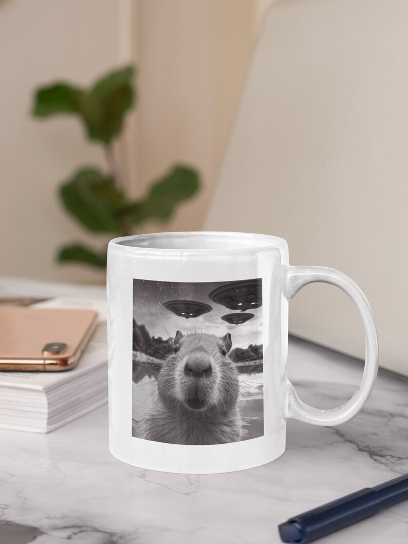 Pu&ograve; includere: Una tazza in ceramica bianca con un'immagine in bianco e nero di un capibara che guarda direttamente la fotocamera. Il capibara &egrave; in un specchio d'acqua con tre dischi volanti nel cielo sopra.