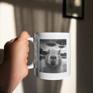 Pu&ograve; includere: Una tazza in ceramica bianca con un'immagine in bianco e nero di un capibara con tre UFO sullo sfondo.