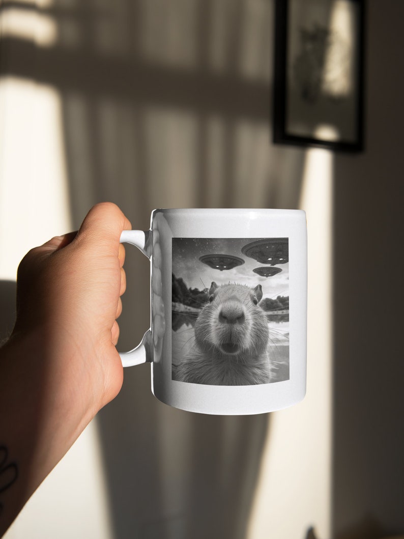 Pu&ograve; includere: Una tazza in ceramica bianca con un'immagine in bianco e nero di un capibara con tre UFO sullo sfondo.