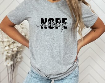 Könnte beinhalten: Hellgraues T-Shirt mit dem schwarzen Aufdruck "NOPE not today". Das Wort "NOPE" ist fett und ohne Serifen, "not today" ist kursiv geschrieben. Das T-Shirt ist aus einem weichen Material gefertigt.
