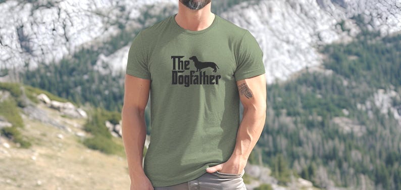 May include: An olive green t-shirt featuring the words "The Dogfather" and a dachshund dog silhouette. The t-shirt is made of a soft material and is suitable for casual wear. A great gift for dog lovers.
