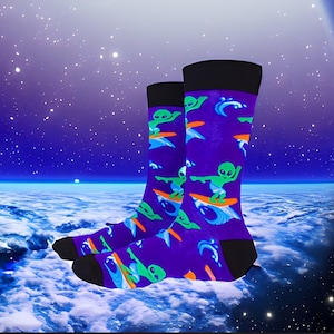 May include: A pair of purple socks with a pattern of green aliens surfing on orange surfboards against a blue and white wave background.