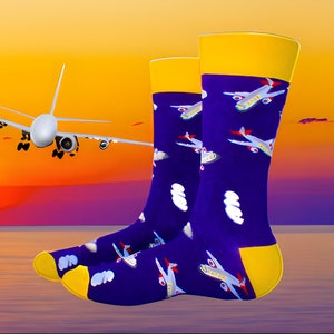May include: A pair of purple socks with a yellow cuff and a pattern of white clouds and airplanes. The socks are photographed against a sunset background with a white airplane flying in the distance.