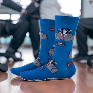 May include: A pair of blue socks with a cartoon chimpanzee wearing a suit and tie. The socks have a white sole and a red and white design. The background is blurred, showing office chairs and people.