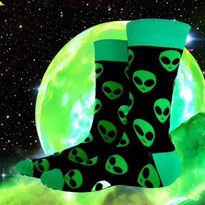 May include: A pair of black socks with a green alien face pattern against a backdrop of a green planet and a crescent moon in a starry sky.