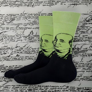 May include: A pair of black and green socks with a portrait of a man on each sock. The socks are on a white background with black ink markings.