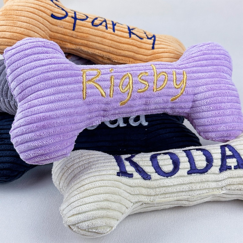 K&ouml;nnte beinhalten: Ein Stapel aus vier knochenf&ouml;rmigen Hundespielzeugen aus Kordstoff. Das oberste Spielzeug ist hellbraun mit blauem Schriftzug Sparky. Das zweite ist lavendelfarben mit goldenem Schriftzug Rigsby. Das dritte ist dunkelblau und das unterste ist cremefarben mit blauem Schriftzug KODA.