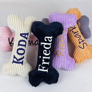 K&ouml;nnte beinhalten: Eine Sammlung von Pl&uuml;sch-Hundespielzeug in Knochenform in verschiedenen Farben, darunter Rosa, Creme, Marineblau, Lila und Orange. Jeder Knochen hat einen gestickten Namen in einer Kontrastfarbe. Die Spielzeuge haben eine Cordstruktur.