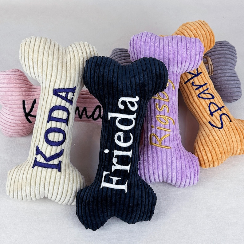 K&ouml;nnte beinhalten: Eine Sammlung von Pl&uuml;sch-Hundespielzeug in Knochenform in verschiedenen Farben, darunter Rosa, Creme, Marineblau, Lila und Orange. Jeder Knochen hat einen gestickten Namen in einer Kontrastfarbe. Die Spielzeuge haben eine Cordstruktur.