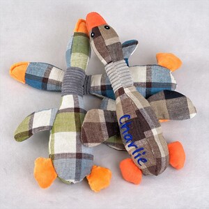 May include: Three soft goose toys with a plaid design in brown, green, blue, and white. The geese have orange beaks and feet, grey necks, and the name "Charlie" embroidered in blue on one. These toys are designed for pets.