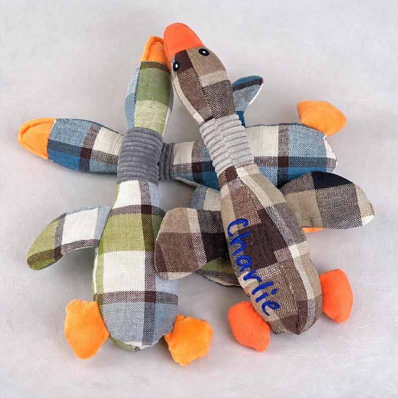 May include: Three soft goose toys with a plaid design in brown, green, blue, and white. The geese have orange beaks and feet, grey necks, and the name "Charlie" embroidered in blue on one. These toys are designed for pets.