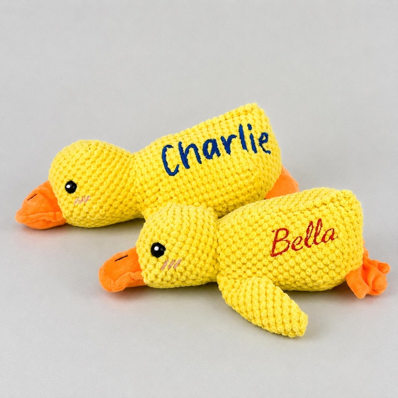 May include: Two yellow plush duck toys with orange beaks and feet. One duck has the name "Charlie" embroidered in blue, and the other has "Bella" in red. The ducks have black eyes and are lying down.