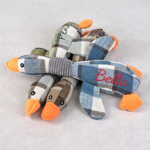 May include: A dog toy in the shape of a duck, made from fabric with a plaid design in blue, green, and brown. The duck has orange feet and beak, with the name "Bella" embroidered in red. The name "Penny" is embroidered in green on another part of the toy.