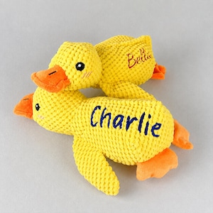 May include: Two yellow plush duck toys with orange beaks and feet. One duck has the name "Charlie" embroidered in blue, and the other has "Bella" embroidered in red. The ducks are made of a textured fabric.