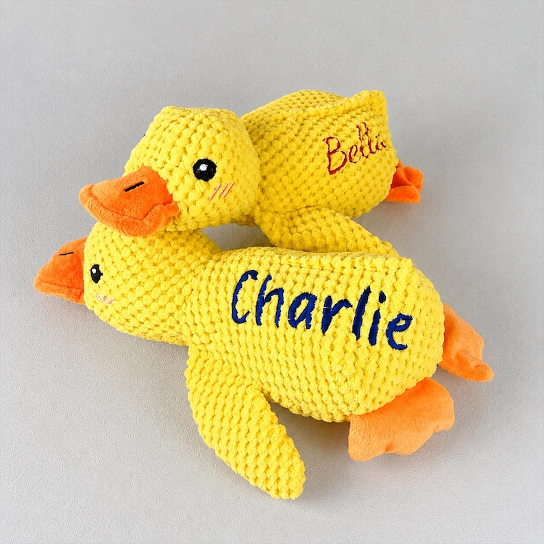 May include: Two yellow plush duck toys with orange beaks and feet. One duck has the name "Charlie" embroidered in blue, and the other has "Bella" embroidered in red. The ducks are made of a textured fabric.