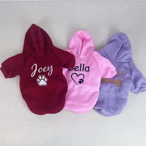 Può includere: Tre felpe con cappuccio per cani in bordeaux, rosa e lavanda. La felpa bordeaux ha "Joey" e un'impronta di zampa. La felpa rosa ha "Bella", un cuore e un'impronta di zampa. La felpa lavanda ha un osso.
