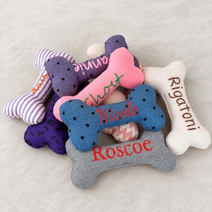 May include: A collection of plush bone-shaped toys in various fabrics including purple stripes, black bat prints, pink, blue polka dots, and cream herringbone. Each toy features embroidered names including Daisy, Annie, Ghost, Nicole, and Roscoe.