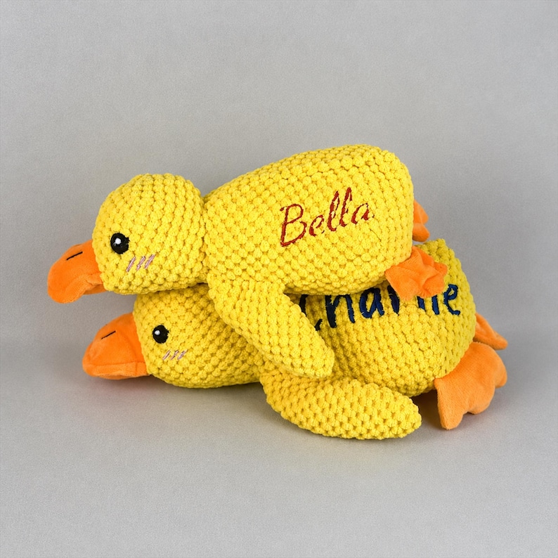 May include: Two yellow knitted duck plush toys, one atop the other. The top duck has the name "Bella" embroidered in red. The bottom duck has the name "Charlie" embroidered in blue. Both ducks have orange beaks and feet, and black eyes.