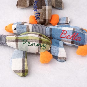 May include: Three soft duck toys with blue, green, and brown plaid patterns. Each duck has orange feet and beak, with names embroidered on them: "Charlie", "Penny", and "Bella". The toys have grey ribbed necks.