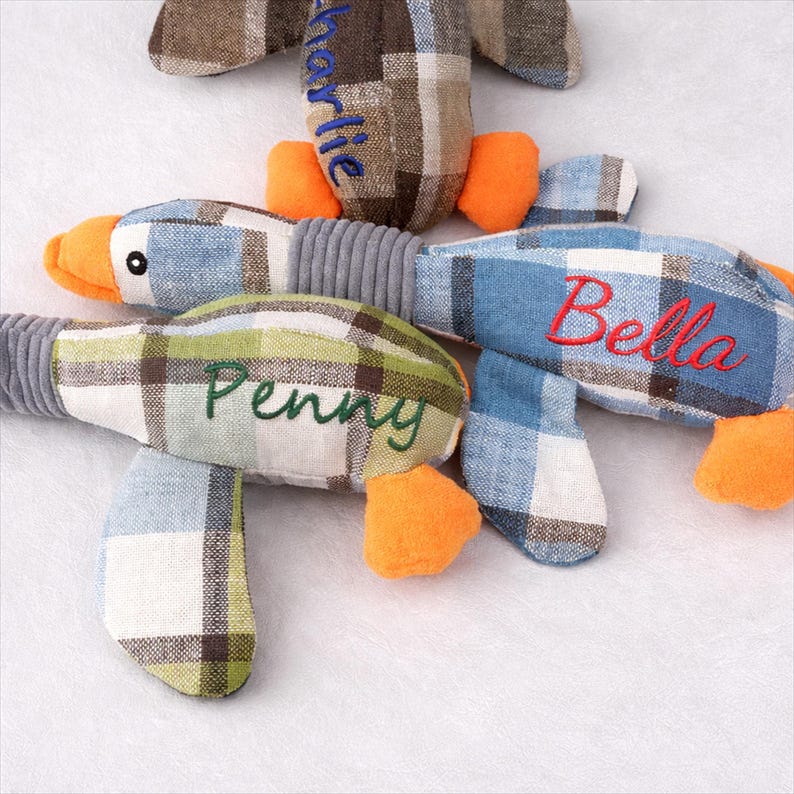 May include: Three soft duck toys with blue, green, and brown plaid patterns. Each duck has orange feet and beak, with names embroidered on them: "Charlie", "Penny", and "Bella". The toys have grey ribbed necks.