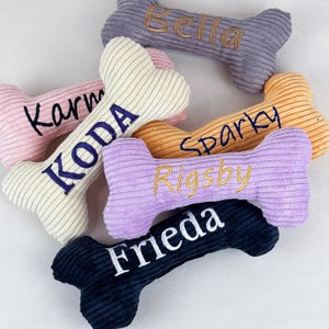 K&ouml;nnte beinhalten: Eine Sammlung von sechs knochenf&ouml;rmigen Cord-Pl&uuml;schtieren in verschiedenen Farben wie Grau, Rosa, Creme, Orange, Lavendel und Marineblau. Jedes Spielzeug ist mit den Namen Bella, Karma, KODA, Sparky, Rigsby und Frieda in Kontrastgarn bestickt.