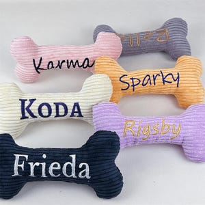 K&ouml;nnte beinhalten: Eine Sammlung von sechs knochenf&ouml;rmigen Hundespielzeugen aus Kord in verschiedenen Farben, jeweils mit einem Namen bestickt: Karma in Schwarz, Bella in Braun, Sparky in Blau, Koda in Dunkelblau, Rigsby in Gold und Frieda in Wei&szlig;.
