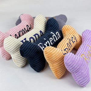 K&ouml;nnte beinhalten: Eine Sammlung von knochenf&ouml;rmigen Cord-Pl&uuml;schtieren in verschiedenen Farben wie Rosa, Creme, Marineblau, Orange und Lavendel. Jedes Spielzeug ist mit gestickten Namen in Schreibschrift versehen, namentlich Karma, Yoda, Frieda, Darky und Rosi.