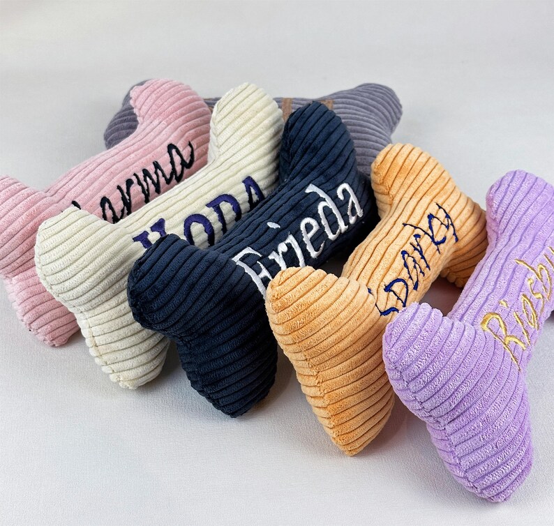 K&ouml;nnte beinhalten: Eine Sammlung von knochenf&ouml;rmigen Cord-Pl&uuml;schtieren in verschiedenen Farben wie Rosa, Creme, Marineblau, Orange und Lavendel. Jedes Spielzeug ist mit gestickten Namen in Schreibschrift versehen, namentlich Karma, Yoda, Frieda, Darky und Rosi.