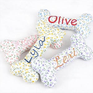 Può includere: Quattro giocattoli a forma di osso per cani con motivi floreali. Ogni osso ha un nome ricamato: "Olive" in rosso, "Lyla" in blu, "Lexi" in arancione e un altro in rosa. Sfondo bianco con piccoli fiori.