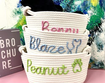Könnte beinhalten: Drei weiße Korbgeflechte aus Seil, übereinander gestapelt. Die Körbe sind mit den Namen "Peanut", "Blaze" und "Remmi" in grünem, blauem und rosa Garn bestickt. Jeder Name wird von einem kleinen gestickten Design begleitet.