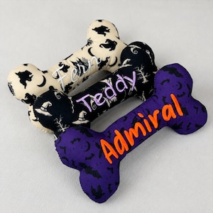 May include: Three bone-shaped plush toys featuring Halloween patterns. The top toy is cream with black ghosts and moons, embroidered with "Fern". The middle is black with witch hats, embroidered with "Teddy". The bottom is purple with bats, embroidered with "Admiral".