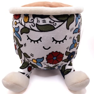 Op de afbeelding: Een pluche koffiekopje speelgoed met een lachend gezicht en gesloten ogen. De beker is wit met een bloemenontwerp in rood, blauw en geel. De bovenkant heeft een bruine, koffie-achtige vulling en een crèmekleurige rand. Het speelgoed heeft kleine, crèmekleurige voetjes.