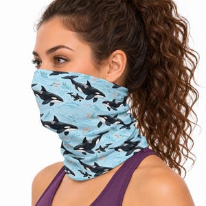 May include: A light blue neck gaiter featuring a repeating pattern of black and white orca whales swimming among small orange fish and green seaweed illustrations. The fabric is lightweight and covers the lower face and neck area.