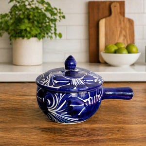 May include: A cobalt blue ceramic casserole dish with a lid, featuring a white floral design. The dish has a handle and a rounded shape, perfect for serving. The background includes a wooden surface and a white countertop.