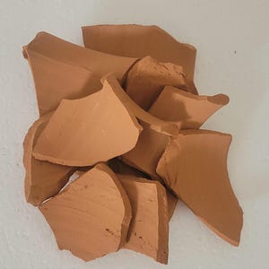 Op de afbeelding: Een stapel gebroken terracotta aardewerkfragmenten. De stukken, in verschillende maten en vormen, vertonen een warme, aardse bruine kleur. De gebroken randen tonen de textuur en interne structuur van het materiaal.