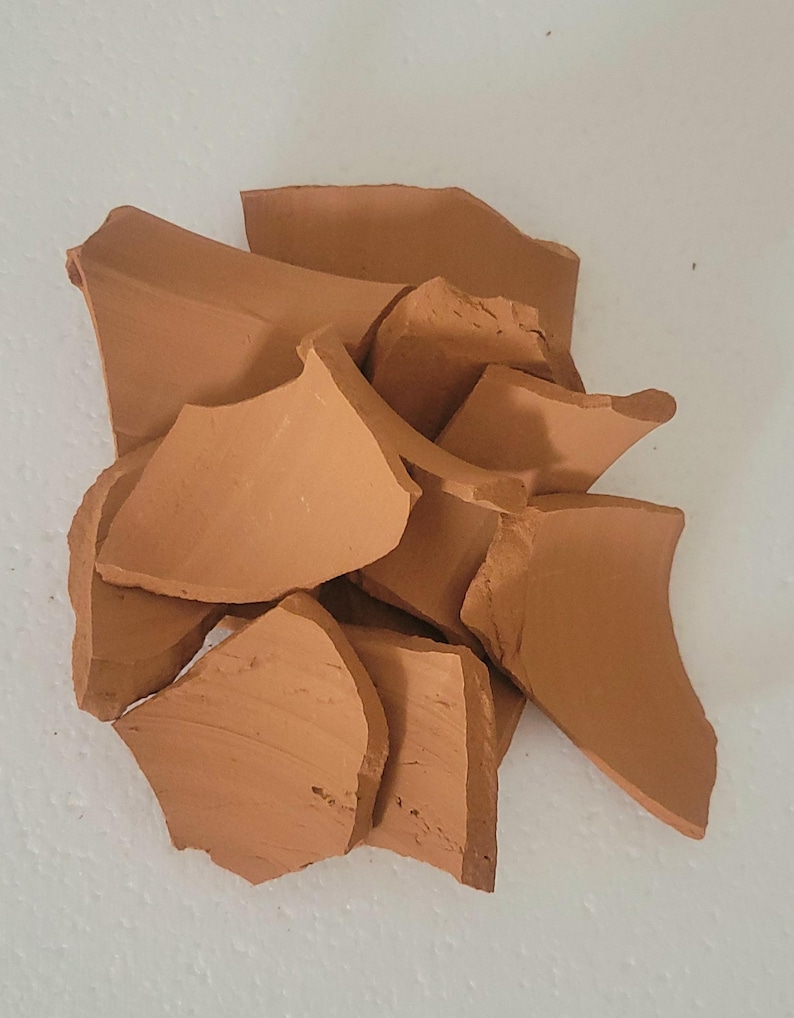 Op de afbeelding: Een stapel gebroken terracotta aardewerkfragmenten. De stukken, in verschillende maten en vormen, vertonen een warme, aardse bruine kleur. De gebroken randen tonen de textuur en interne structuur van het materiaal.
