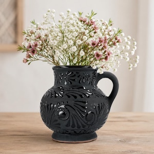 May include: A black ceramic vase with a handle, filled with white and pink flowers, sits on a wooden table. The vase has a detailed, embossed floral design. The background includes a window and a decorative wooden frame.