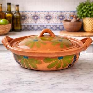 May include: A terracotta cooking pot with a lid, featuring a floral design in green and blue. The pot has two handles and a loop on the lid. It is sitting on a marble countertop.