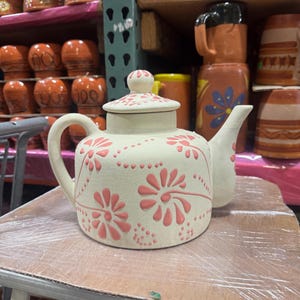 May include: Cream-colored ceramic teapot with a lid and a curved spout. The teapot is decorated with pink floral designs and small dots. The handle is curved and the teapot is sitting on a table.