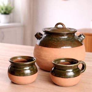 May include: A set of three handcrafted ceramic pieces: a large pot with a lid and two smaller vessels. The pottery features a two-tone design with a natural terracotta base and a dark green glazed upper section. The pot has two handles.
