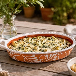 May include: An oval, orange ceramic serving dish filled with a baked dip, with white handles and a decorative white floral design. A glass of white wine, a basket of tortilla chips, and a bowl of bread slices are also present.