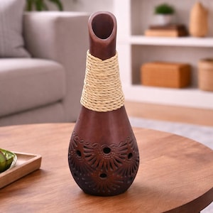 May include: A dark brown decorative vase with a narrow neck and a wide base. The vase has a woven tan band around the neck and intricate floral cutouts near the base. It sits on a round wooden table.