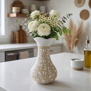 May include: A ceramic vase with a beige base and white floral design, holding a bouquet of white flowers and greenery. The vase has a white, scalloped rim. The arrangement sits on a white countertop in a kitchen setting.