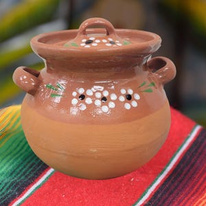 May include: A handcrafted, round, terracotta clay pot with a lid. The pot features two handles and is decorated with white floral designs and green leaves. The pot is sitting on a colorful, striped textile.
