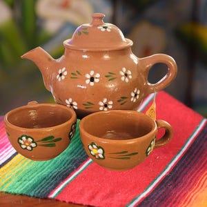 May include: A terracotta tea set featuring a teapot with a lid and two teacups. The set is decorated with white and green floral designs. The tea set is displayed on a colorful striped textile.