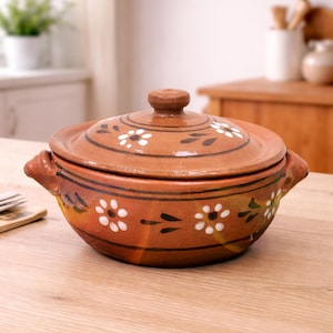 May include: A terracotta-colored ceramic casserole dish with a lid. The dish features white floral designs and dark brown accents. It has two handles and sits on a wooden surface, suggesting a rustic kitchenware theme.